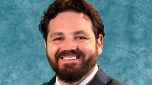 Smiling man with a beard in a blue suit and tie against a mottled teal background. The mood is positive and professional.