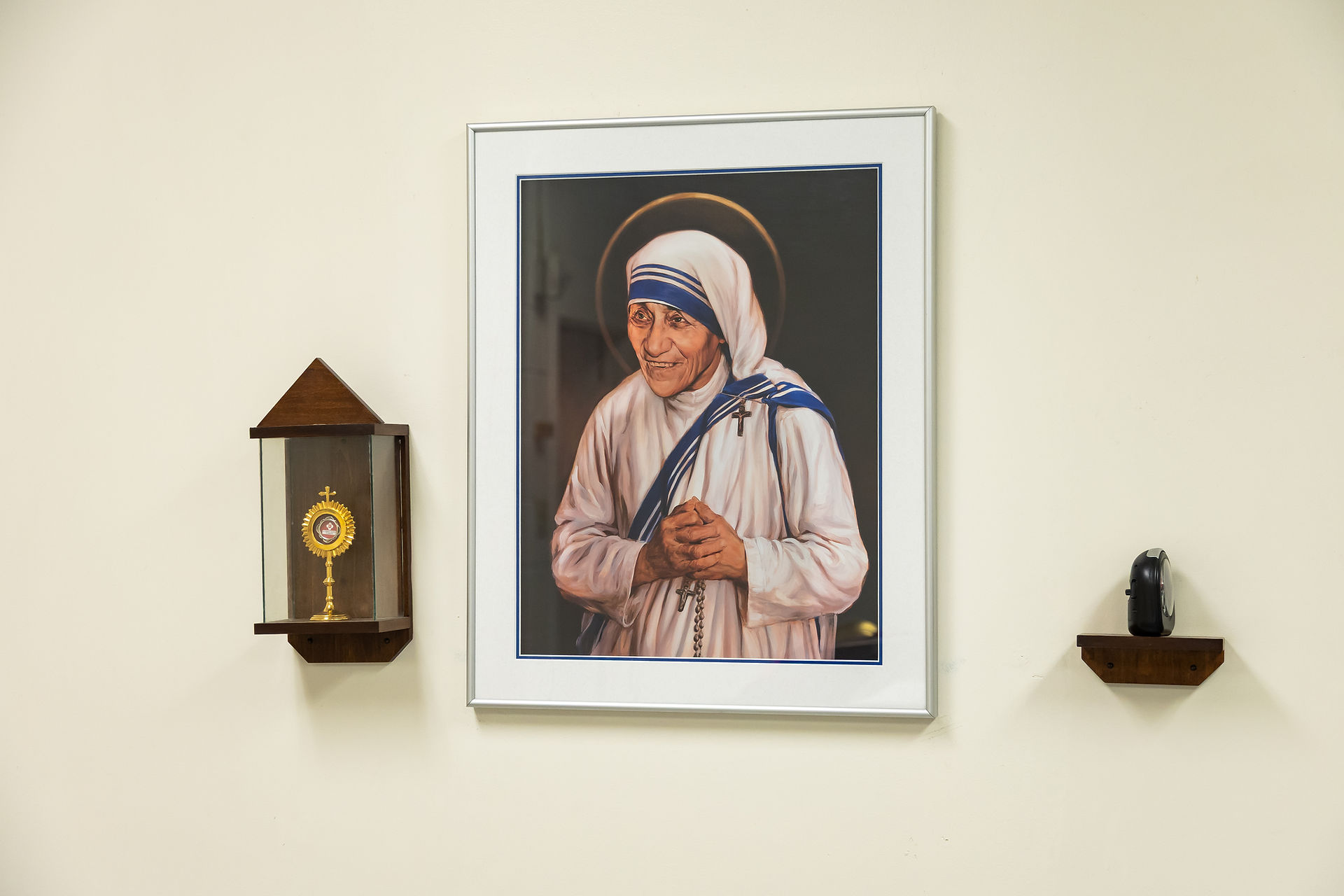 Framed portrait of a smiling nun with a halo on a white wall, flanked by a reliquary and a small object on wooden shelves.