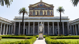 Sunny basilica courtyard with palm trees, statues, and a painted church facade above trimmed green hedges.