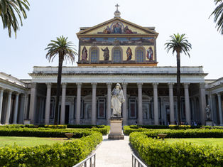 Sunny basilica courtyard with palm trees, statues, and a painted church facade above trimmed green hedges.