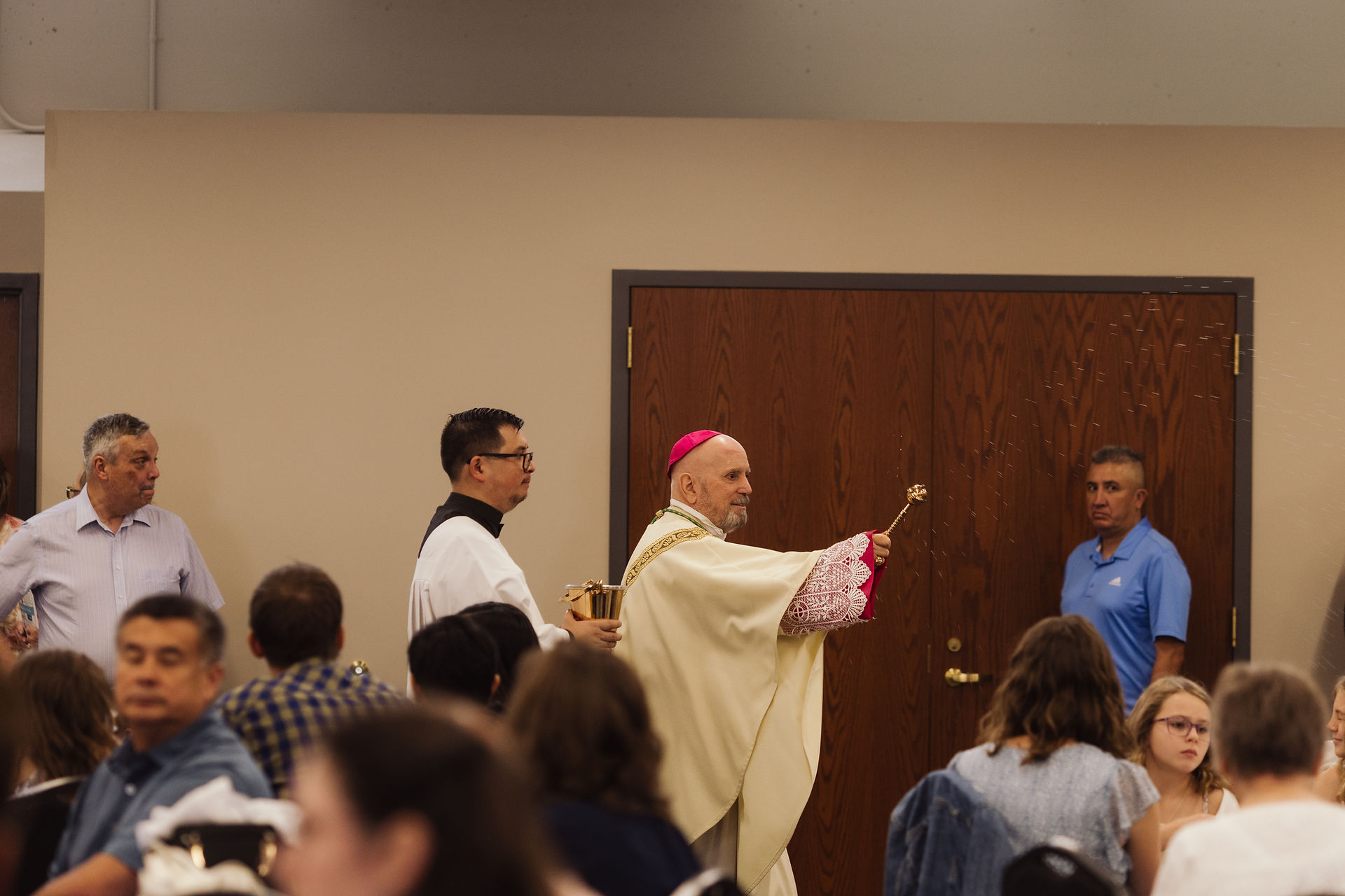 A bishop in cream robes, holding a sprinkler, blesses a room of seated people. A man assists, holding a gold container. Mood is solemn.