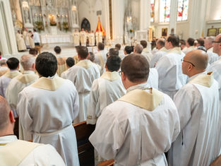 Priests in white robes with gold stoles gather in a cathedral during a ceremony. The altar, candles, and stained glass are visible.