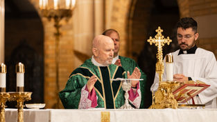 Bishop in green robe raises hands in prayer at altar with gold cross and candles, while another person in white robe looks on. Church setting.