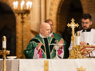 Bishop in green robe raises hands in prayer at altar with gold cross and candles, while another person in white robe looks on. Church setting.