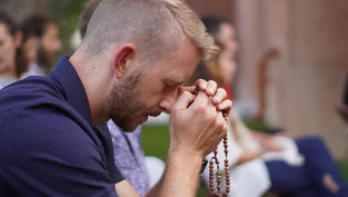 A man prays outdoors, eyes closed, holding rosary beads. Others sit blurred in the background. The mood is calm and reflective.