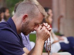 A man prays outdoors, eyes closed, holding rosary beads. Others sit blurred in the background. The mood is calm and reflective.
