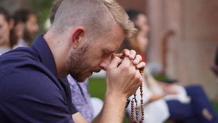 A man prays outdoors, eyes closed, holding rosary beads. Others sit blurred in the background. The mood is calm and reflective.