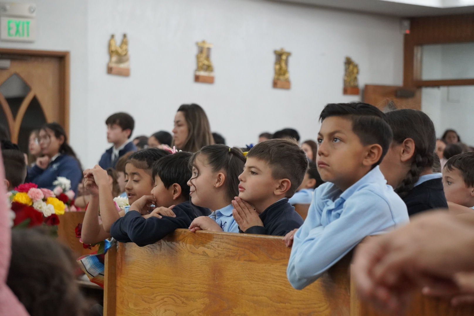 Children attentively listening in a church, resting on wooden pews. Colorful flowers are visible, creating a peaceful and focused mood.