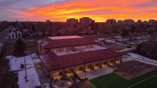 A building with a red roof stands against a vibrant orange sunset, with snow patches and parked cars nearby in an urban setting.