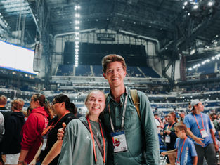 Smiling man and woman at a large indoor event with a crowd and bright lights in the stadium. They wear lanyards and casual clothes. Energetic mood.