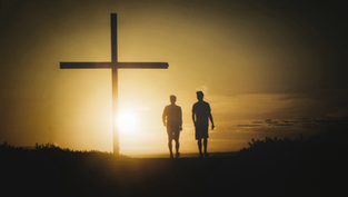 Silhouetted against a golden sunset, two people walk near a large cross on a hilltop, creating a serene and reflective scene.