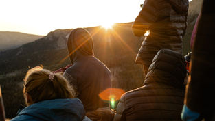 People in jackets watch a sunrise over mountains, creating a warm glow. The scene is peaceful with soft light and natural beauty.