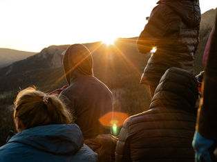 People in jackets watch a sunrise over mountains, creating a warm glow. The scene is peaceful with soft light and natural beauty.