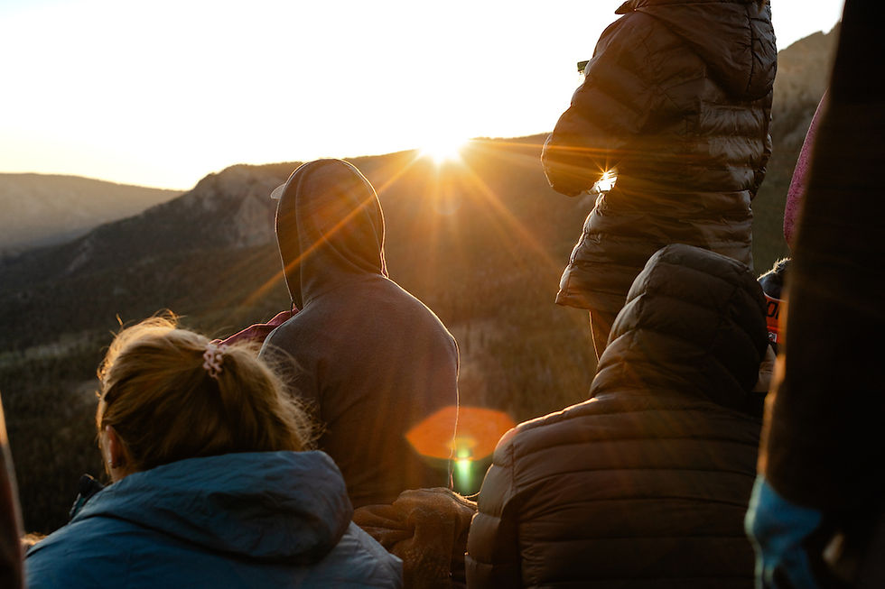 People in jackets watch a sunrise over mountains, creating a warm glow. The scene is peaceful with soft light and natural beauty.