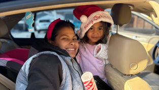 Smiling woman and child in car. Child wears a Santa hat with "Merry Christmas" text. Bright day, car seat visible, happy vibe.