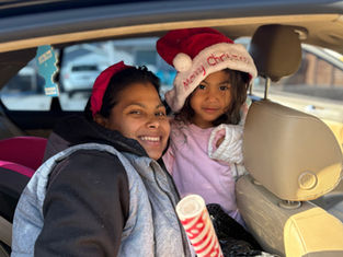 Smiling woman and child in car. Child wears a Santa hat with "Merry Christmas" text. Bright day, car seat visible, happy vibe.