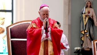 Bishop in red robes prays with eyes closed in a church, next to a statue of a saint. Warm light, reverent mood, stained glass visible.