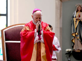 Bishop in red robes prays with eyes closed in a church, next to a statue of a saint. Warm light, reverent mood, stained glass visible.