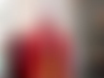 Bishop in red robes prays with eyes closed in a church, next to a statue of a saint. Warm light, reverent mood, stained glass visible.