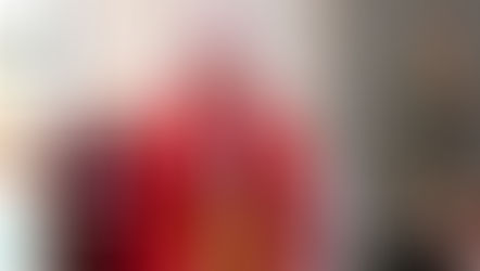 Bishop in red robes prays with eyes closed in a church, next to a statue of a saint. Warm light, reverent mood, stained glass visible.