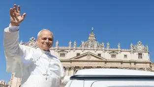 A person in white religious attire waves while standing near a vehicle. The background shows an ornate building with a clear blue sky.