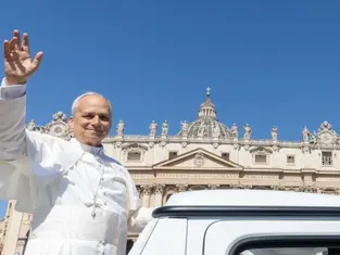 A person in white religious attire waves while standing near a vehicle. The background shows an ornate building with a clear blue sky.