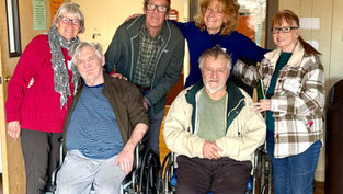 Group of smiling people, including two in wheelchairs, pose in a warmly lit room. One woman holds a green box. Casual, supportive mood.