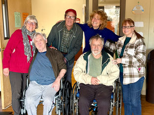 Group of smiling people, including two in wheelchairs, pose in a warmly lit room. One woman holds a green box. Casual, supportive mood.