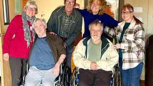 Group of smiling people, including two in wheelchairs, pose in a warmly lit room. One woman holds a green box. Casual, supportive mood.