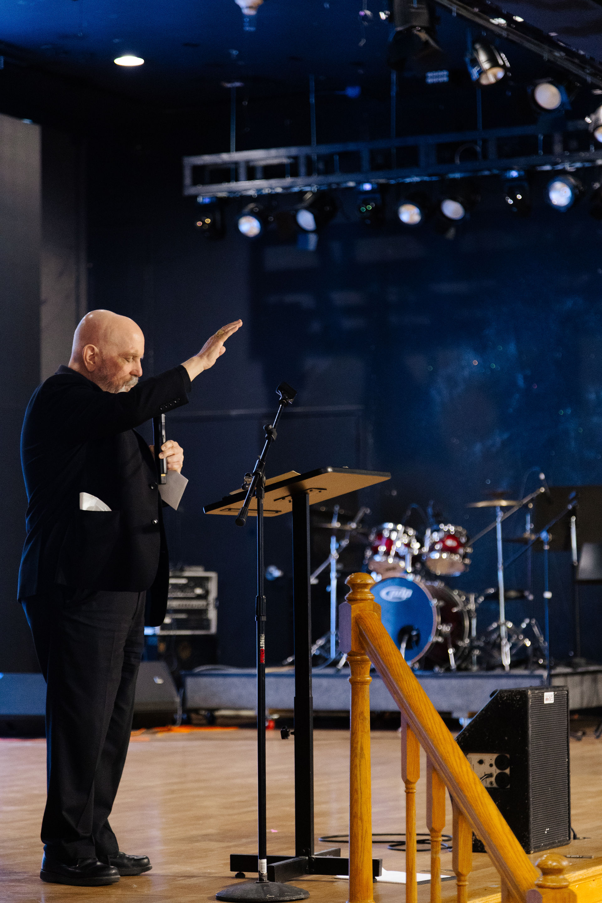 Man stands on a stage with his hand raised in prayer while holding a microphone near a lectern, with musical instruments in the background.