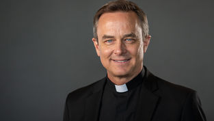 Clergy member wearing a black clerical suit smiles against a plain gray background, exuding a warm and welcoming demeanor.