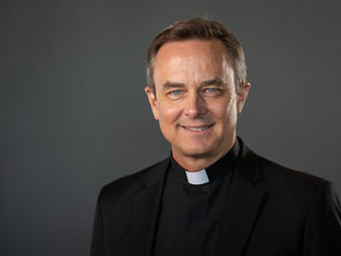 Clergy member wearing a black clerical suit smiles against a plain gray background, exuding a warm and welcoming demeanor.
