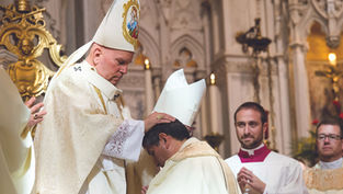 A bishop in ornate vestments places hands on a kneeling man in a church ceremony. Two clergy members observe. Emotive, ceremonial setting.
