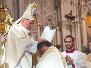 A bishop in ornate vestments places hands on a kneeling man in a church ceremony. Two clergy members observe. Emotive, ceremonial setting.