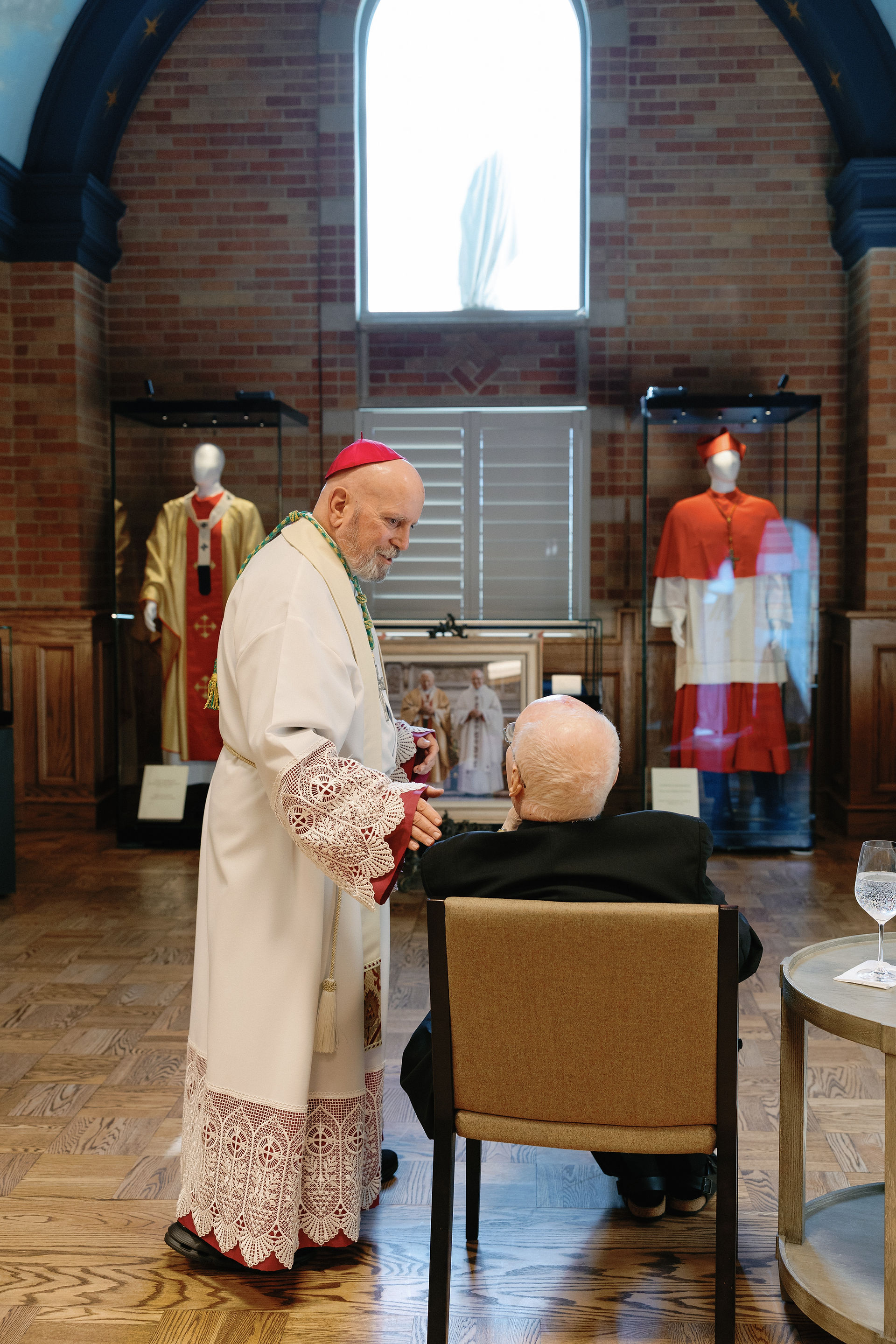 A bishop in ornate robes speaks to a seated man in a room with religious garments displayed. The mood is solemn and respectful.