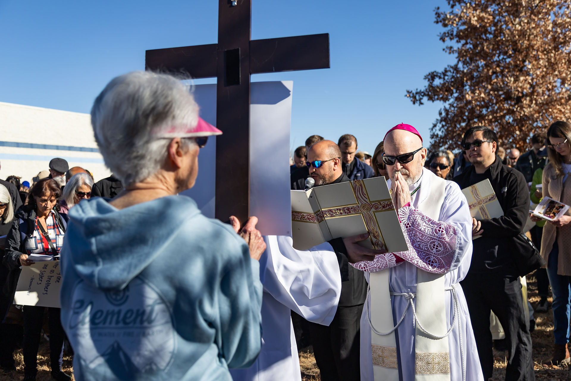 Clergy and crowd gather outdoors for religious event, holding books and a large cross. Blue sky, brown leaves on tree, solemn mood.
