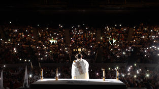 A priest in white holds a monstrance on stage. Candles lit on an altar. Audience in dim light with phone lights, creating a reverent mood.
