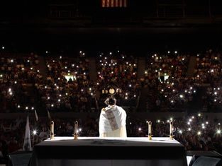 A priest in white holds a monstrance on stage. Candles lit on an altar. Audience in dim light with phone lights, creating a reverent mood.