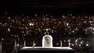 A priest in white holds a monstrance on stage. Candles lit on an altar. Audience in dim light with phone lights, creating a reverent mood.