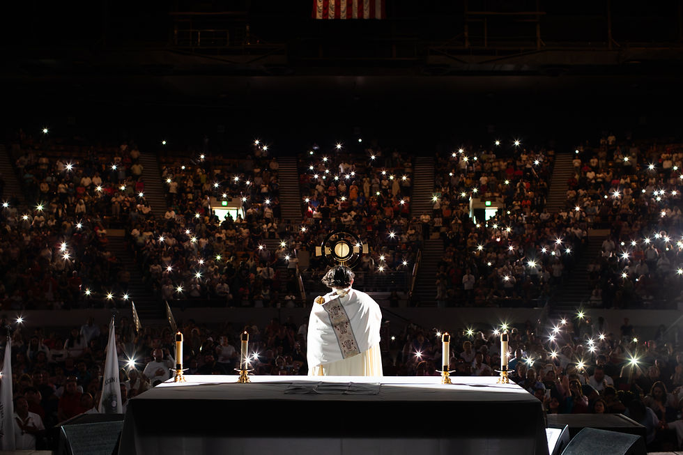 A priest in white holds a monstrance on stage. Candles lit on an altar. Audience in dim light with phone lights, creating a reverent mood.