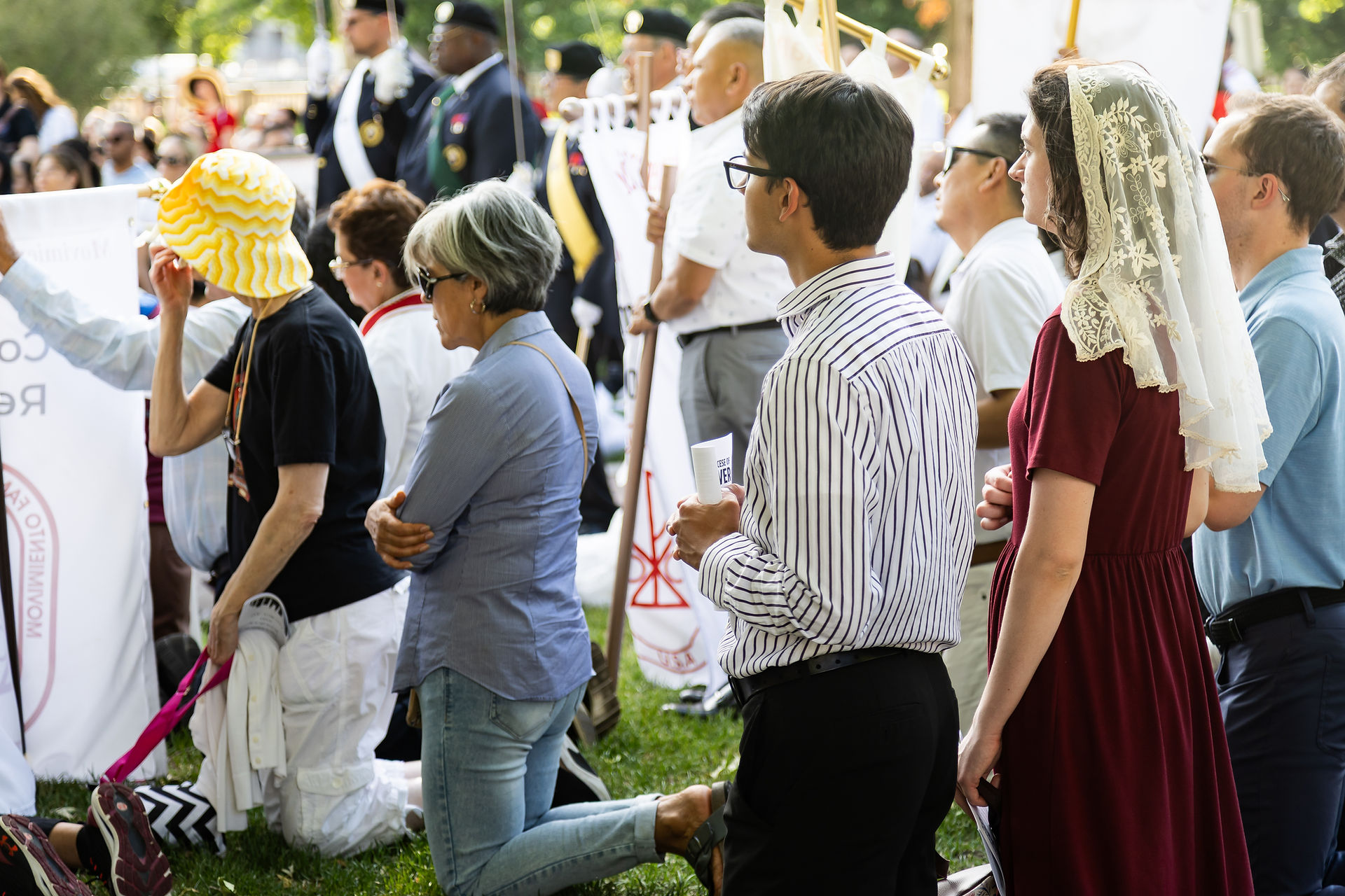 People kneel at an outdoor religious vigil, holding white banners; a woman in a lace veil stands beside them.