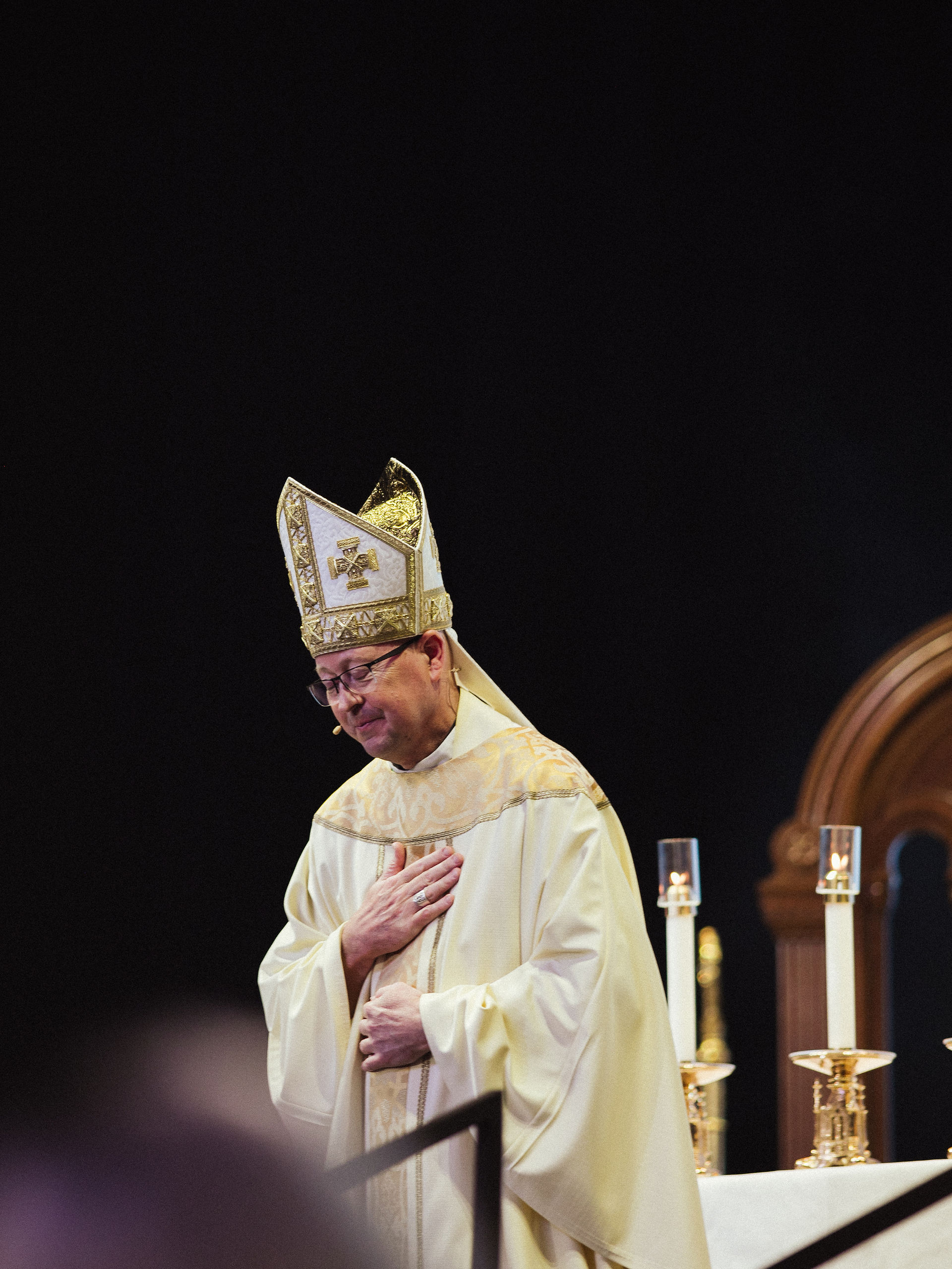 Bishop in ornate robes and mitre stands solemnly at an altar with two lit candles, hand on chest, against a dark background.