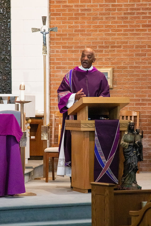 A deacon in a purple robe speaks at a wooden pulpit in a brick-walled church, next to a statue. An altar with a cross is visible.