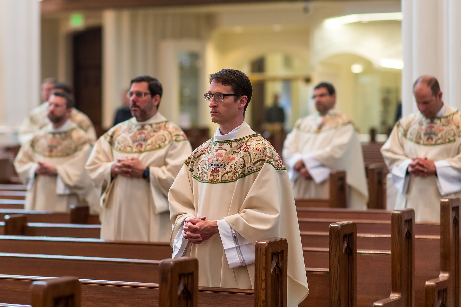 Priests in ornate robes stand with hands clasped in a church, surrounded by wooden pews, conveying a solemn, reflective mood.