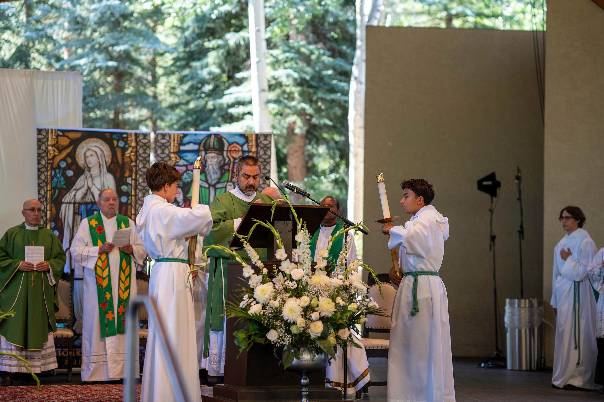 Deacon in green robes reads at a podium with altar servers holding candles. Stained glass backdrop, white flowers, outdoor setting.