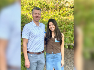 Man and woman smiling outdoors, man in patterned shirt, woman in brown vest with blue skirt. Sunlit leafy background. Both wear name tags.
