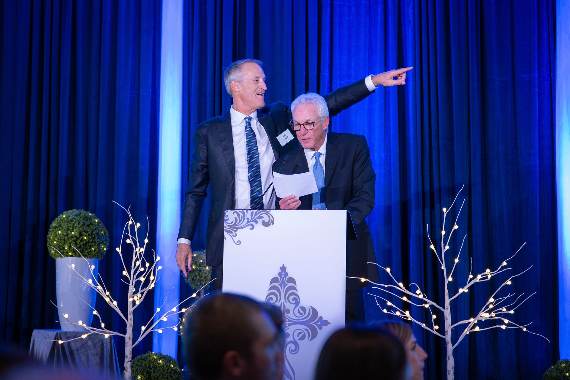 Two men in suits stand at a podium with a decorative panel. One reads a paper, the other points. Blue curtains and lit branches adorn the stage.