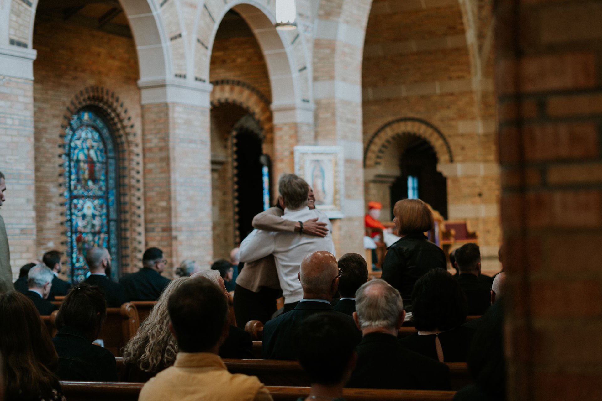People in a church with stained glass, two hugging, others seated, creating a warm, reflective atmosphere. Brick arches in the background.