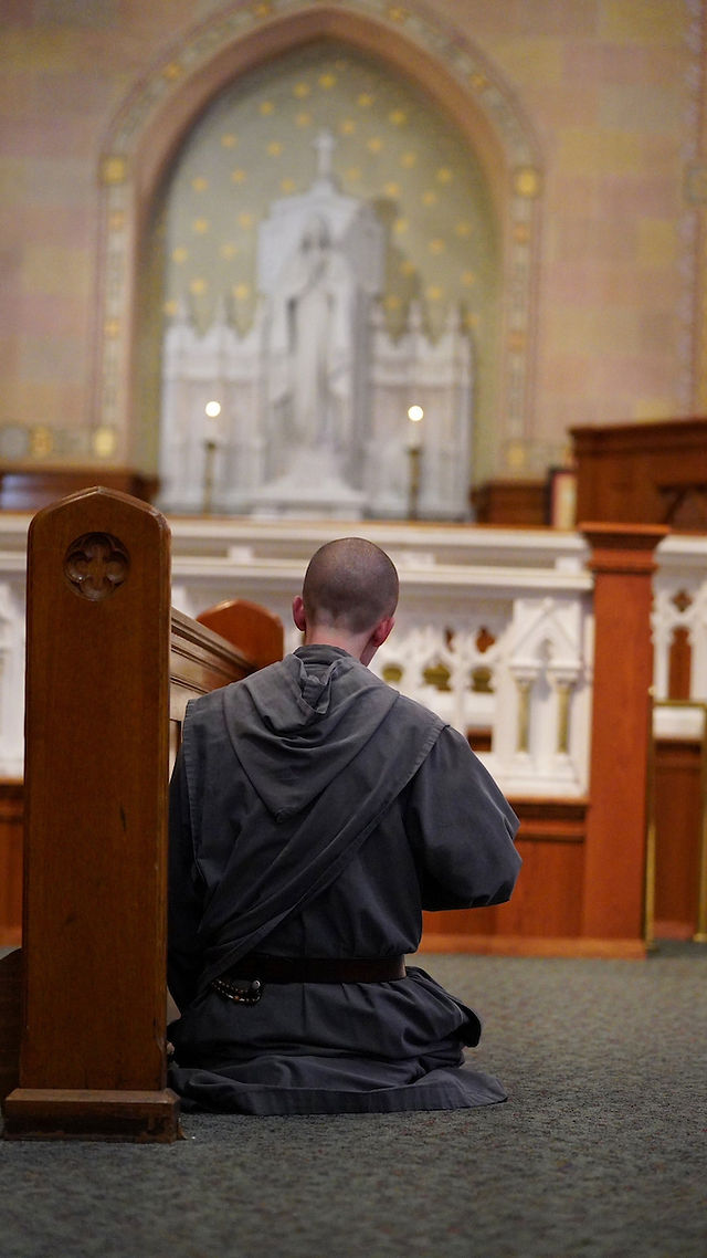 A person in a gray robe kneels in prayer at a church altar, with a stone sculpture in the background. The mood is serene and reflective.