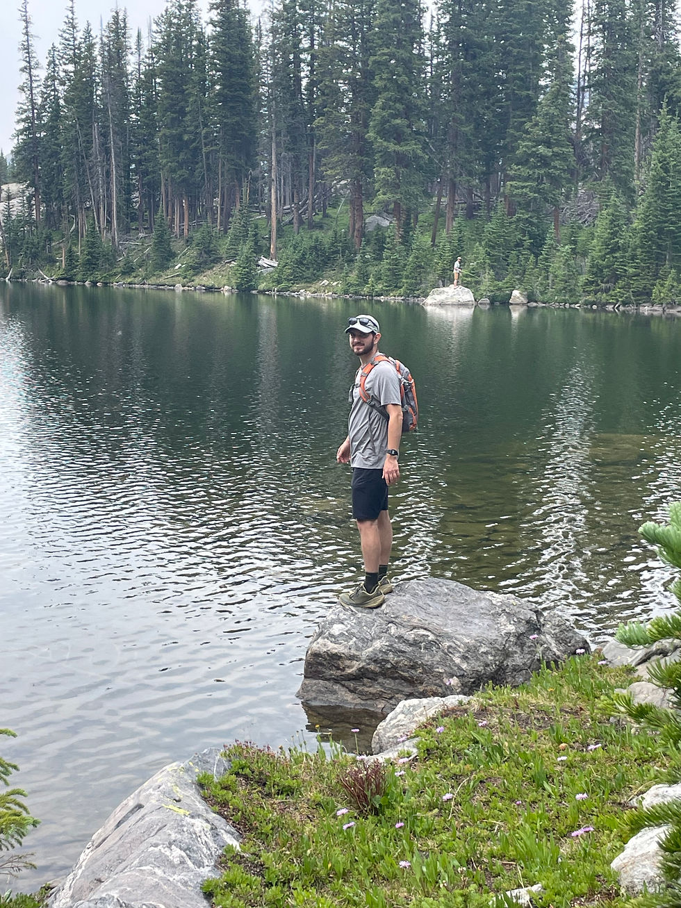 Hiker with backpack stands on a rock by a calm forest lake, with tall pine trees and another person on a distant boulder.